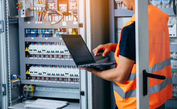 Technicien spécialisé en maintenance industrielle travaillant sur une armoire TGBT avec un ordinateur pour établir un rapport.